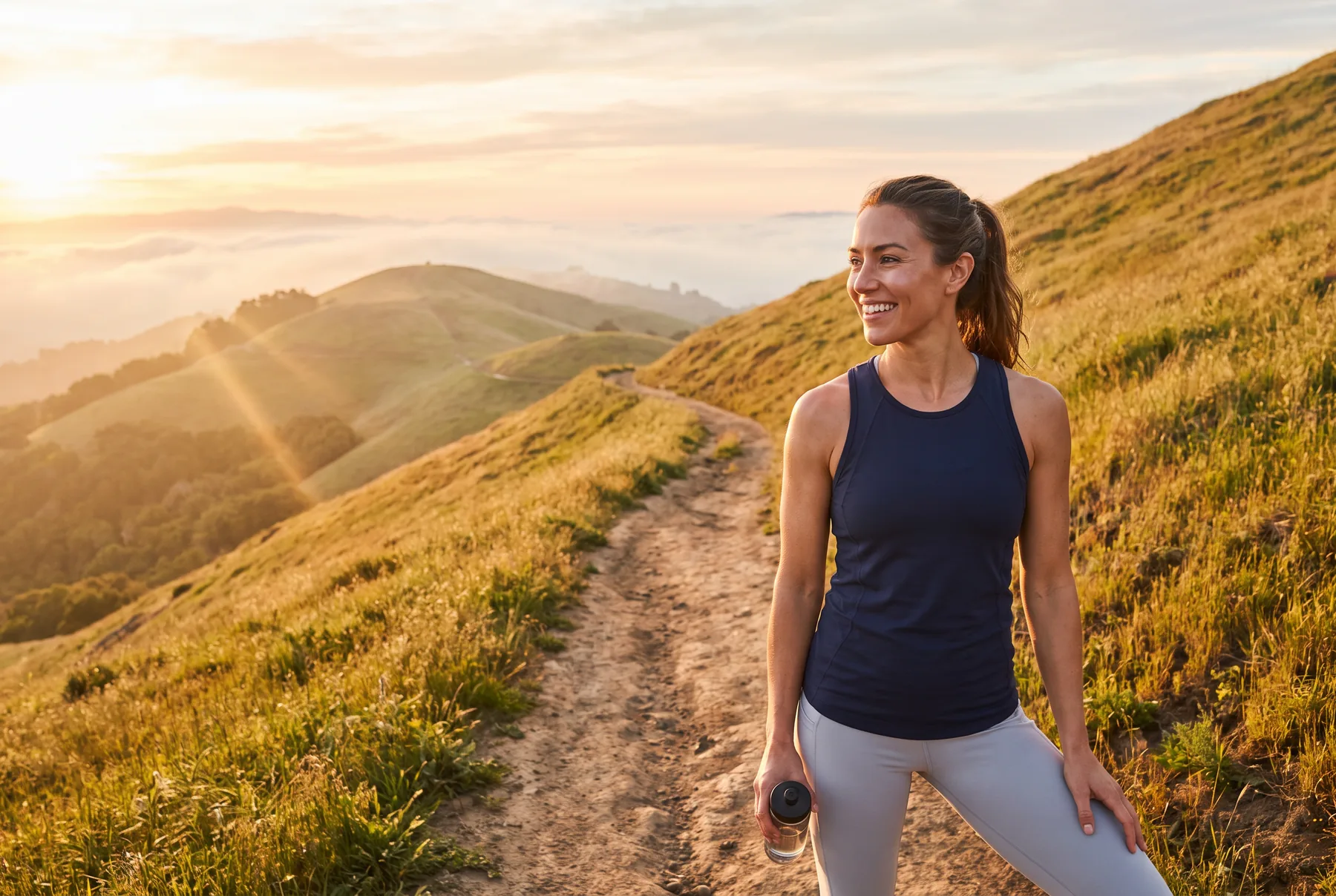 Fit woman smiling on a hillside trail at sunrise