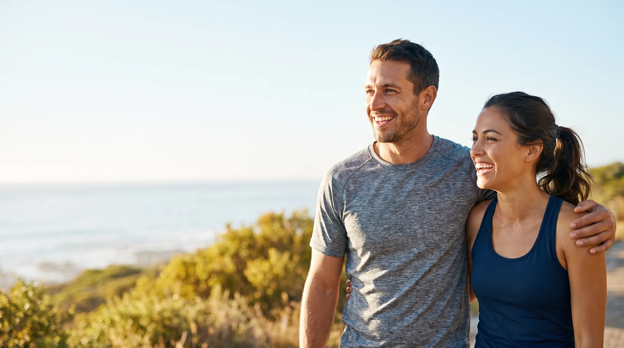 Pareja sonriente con ropa de ejercicio caminando junta por un sendero costero soleado